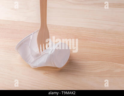 White plastic cup crumpled with a wooden eco fork on the wood table. The concept of rejection of plastic utensils and environmental protection Stock Photo