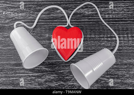 The phone is made of white plastic cups and a white rope laid around a red wooden heart on a black table. The concept of emotional contact between lov Stock Photo