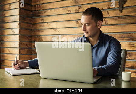 Dedicated businessman taking notes in a journal as he sits at his desk in the office working on a laptop computer in a close up view Stock Photo