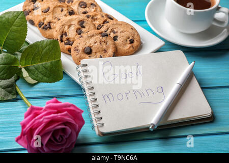 Plate with tasty chocolate chip cookies, cup of coffee and GOOD MORNING note in notebook on color wooden table Stock Photo