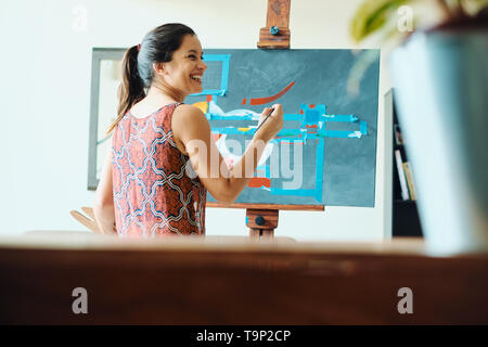 Portrait of hispanic young woman painting in her home studio, exercising with paint, brush, easel and color for hobby. Stock Photo
