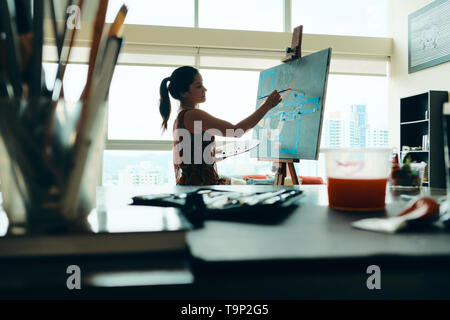 Portrait of young latino woman painting for hobby in her home studio, practicing with paint, brushes, easel and colors. Stock Photo