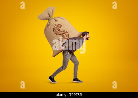 Side view of young handsome man in casual clothes carrying huge heavy sack with dollar symbol on. Stock Photo