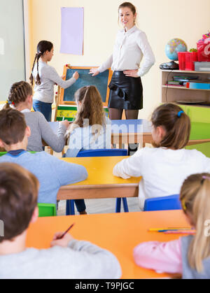 Positive friendly  pleasant female teacher standing at blackboard with schoolgirl during lesson in classroom Stock Photo