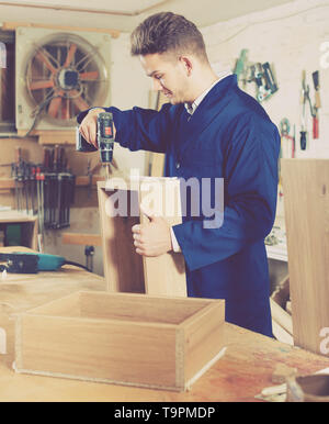Young smiling guy using drilling machine to make drawer at workshop Stock Photo
