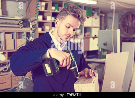 Young smiling joiner using drilling machine to make drawer at workshop Stock Photo