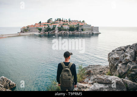 Tourist with a backpack near the sea. Travel alone. Looks into the distance. Ahead is the island of Sveti Stefan in Montenegro. Stock Photo