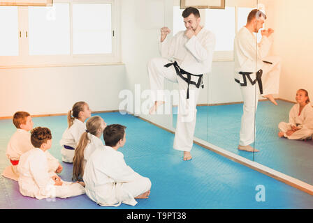 Happy young male coach explaining new maneuvers to children in karate class. Focus on the boy Stock Photo