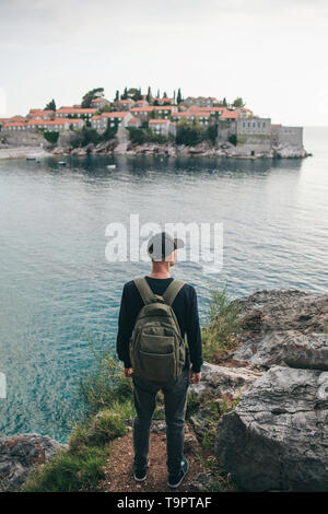 Tourist with a backpack near the sea. Travel alone. Looks into the distance. Ahead is the island of Sveti Stefan in Montenegro. Stock Photo