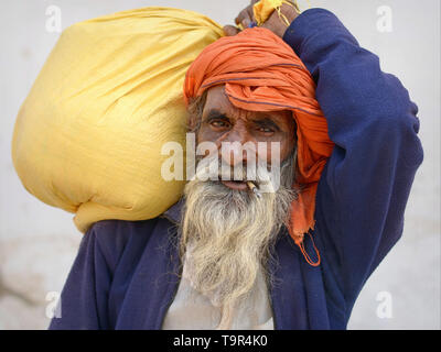 Elderly Indian Rajasthani man smokes a traditional Indian mini-cigar (beedi) and carries a heavy sack on his shoulder. Stock Photo