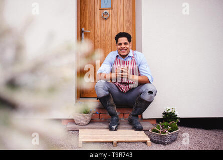 A portrait of young man gardener sitting in front of door at home. Stock Photo