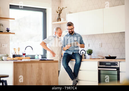 Adult hipster son and senior father indoors in kitchen at home, using tablet. Stock Photo