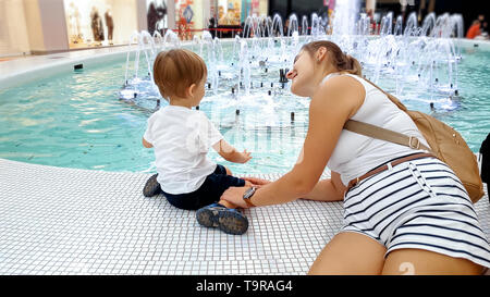 Portrait of happy smiling young mother with little child sitting in shopping mall and looking on the beautiful fountain in hall Stock Photo