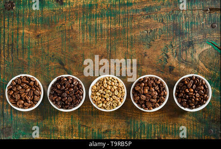 Small bowls of assorted speciality raw, medium and full roast coffee beans on old vintage or rustic wood with copy space arranged as a lower border Stock Photo