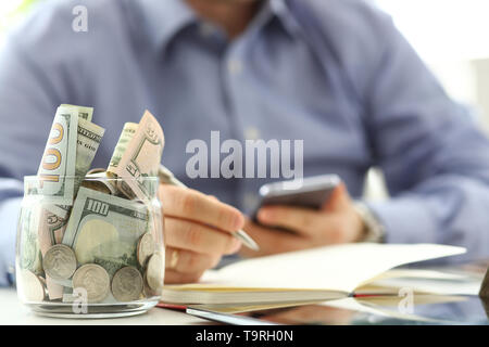 Rich jar full or US banknotes and coins with businessman arms in background counting expenses with his cellphone Stock Photo