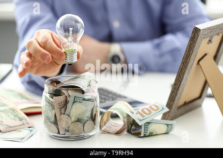 Male arm holding lightbulb over tip can full of US money Stock Photo