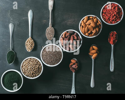 Above view of various superfoods in spoons and small bowl on black wooden table background. Superfood close up chia seeds, spirulina, cocoa bean, goji berry, hemp seeds, almond. Top view, flat-lay Stock Photo