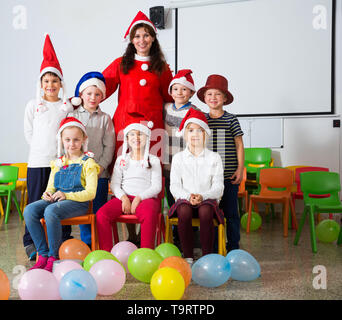 Group of cheerful pupils with female teacher wearing Santa hats posing together in schoolroom Stock Photo