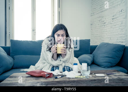 Sick young woman sitting on sofa drinking hot drink cold flu medicine feeling unwell with headache sore nose and high temperature not being able to go