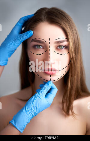 Beautiful young woman with perforation lines on her face before plastic surgery operation. Beautician hands in gloves touching woman face. Puncture Stock Photo