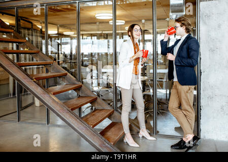 Young buiness man and woman having a conversation standing with cups during the coffee break in the hall of the modern office Stock Photo