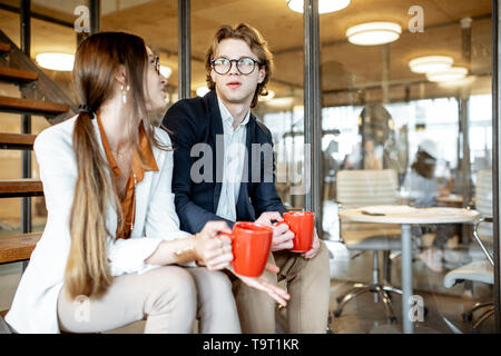 Young buiness man and woman having a conversation, sitting on the stairs during the coffee break in the modern office Stock Photo