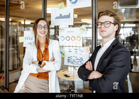Portrait of a business man and woman standing near the charts and drawings on the glass wall in the office Stock Photo