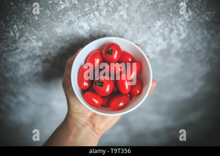 Fresh looking cherry tomatoes in the bowl, holding in hand on gray and white background taken from the top. Stock Photo