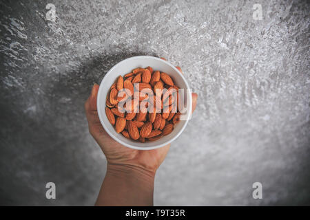 Almonds with skin without shell in the white bowl holding in hand on the gray and white background. Stock Photo