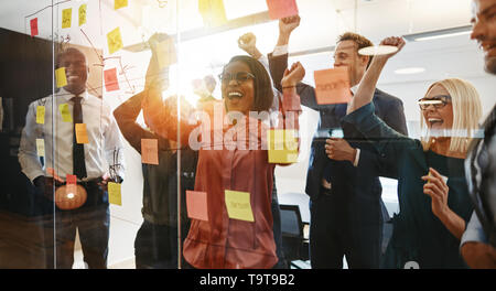 Ecstatic group of diverse businesspeople celebrating a winning idea together while brainstorming with sticky notes on a glass wall in a modern office Stock Photo