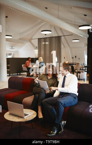 Two diverse coworkers discussing paperwork together while sitting on a sofa in a modern office Stock Photo