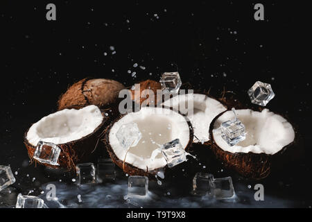 Halves of ripe coconuts, ice cubes and splash of milk on dark background Stock Photo