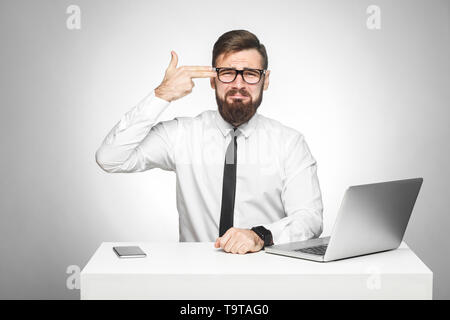 Unsatisfied stressed tired young manager in white shirt and black tie are sitting in office and trying to kill himself showing sign with finger pistol Stock Photo