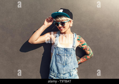 Young alternative girl wearing cap and sunglasses standing isolated on grey wall on the city street smiling cheerful Stock Photo