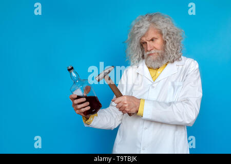 Old long-haired man wearing doctor's gown studio isolated on blue wall standing holding bottle of spirit beating with hammer concerned Stock Photo