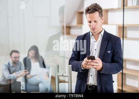 Fashionable business professional at office door using his mobile phone and his colleagues discussing in the background. Stock Photo