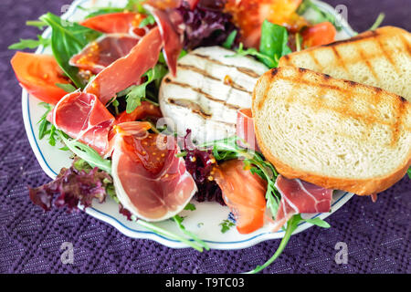 Tasty mixed salad dish with grilled camembert cheese, prosciutto ham, organic tomato and fresh green leaves. Healthy dilicious meal. Isolated on black Stock Photo