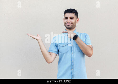 Portrait of happy satisfied handsome young bearded man in blue shirt standing, holding, pointing something and looking at camera with smile. indoor st Stock Photo