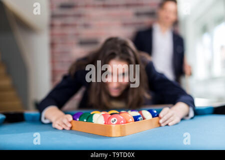 Portrait of young fashionable woman holding triangle wooden rack with pool balls on billiard table. Modern man in the background. Stock Photo