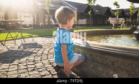 Toned image of adorable little boy sitting next to the fountain on beautfiul old square in park Stock Photo