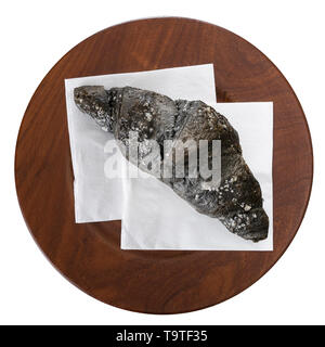 Charcoal croissant, on white serviette and wooden plate, isolated on white. Black colour due to vegetable carbon aka carbo medicinalis vegetabilis. Stock Photo