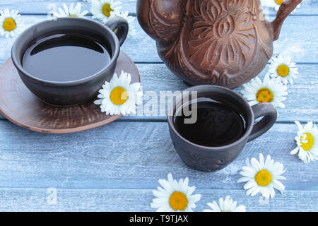 A teapot and a cup of tea stand on wooden blue boards with daisy flowers. Close-up. Stock Photo