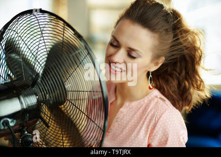 smiling stylish housewife at modern home in sunny hot summer day cooling down in the front of fan. Stock Photo