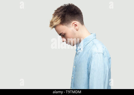 Profile side view portrait of sad upset alone handsome young man in light blue shirt standing, holding head down and depressed. indoor studio shot on  Stock Photo