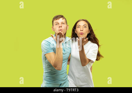Beautiful young couple's half-length portrait isolated on green studio background. Woman and man standing and sending kisses. Facial expression, human emotions concept. Trendy colors. Stock Photo
