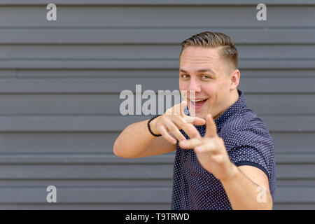 Laughing playful young man gesturing with his hands pointing at the camera in front of a grey wall with copy space Stock Photo