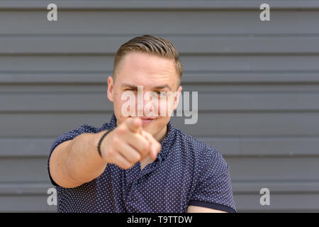 Charismatic young man pointing at the camera with a smile and raised eyebrow posing in front of a grey wall with copy space with focus to his face Stock Photo