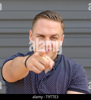 Charismatic young man pointing at the camera with a smile and raised eyebrow posing in front of a grey wall with copy space in square format Stock Photo