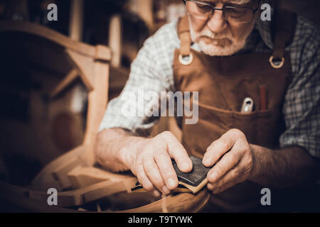 Elderly carpenter with glasses smooths an unfinished chair. workShop in the background. Stock Photo