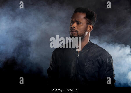Cool strong handsome young african man with beard wearing black windbreaker jacket looking away with serious expression posing against white smoke on  Stock Photo